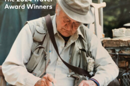 An older white man wearing a hat and fishing vest sits and writes in a notebook using a green pen.