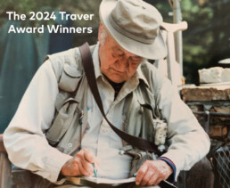 An older white man wearing a hat and fishing vest sits and writes in a notebook using a green pen.