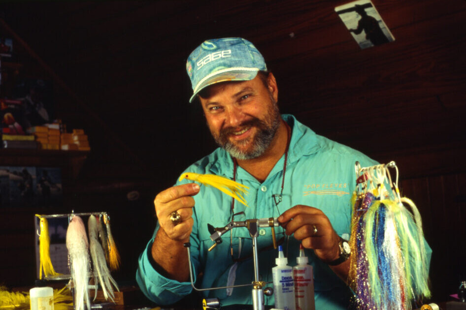 A smiling bearded man sits in front of a fly tying vise, holding up a yellow streamer fly.