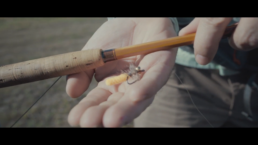 A screenshot featuring a white man holding his right hand out palm up. A bamboo rod is laid across it, the cork handle touching his thumb, and a fly rests along the base of his fingers.