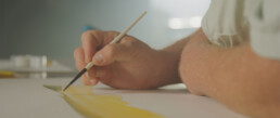 A closeup of a man's hand and arm as he works on a painting of a green and gold fish.