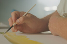 A closeup of a man's hand and arm as he works on a painting of a green and gold fish.