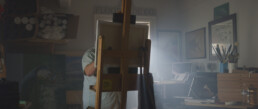 In an artist's work room looking at an easel from behind. All that is visible of the artist is part of his torso and right arm.