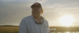 Paul Puckett, a red-haired white man gazes into the distance. He's wearing a black YETI ball cap, a light blue button-down shirt, and sunglasses around his neck. A saltwater marsh in South Carolina's Low Country is the backdrop.