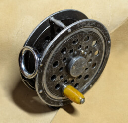 A closeup of a fly reel. It is made of metal that was once coated black, but time and use have revealed much of the gray underneath. The side plate is perforated with several holes.