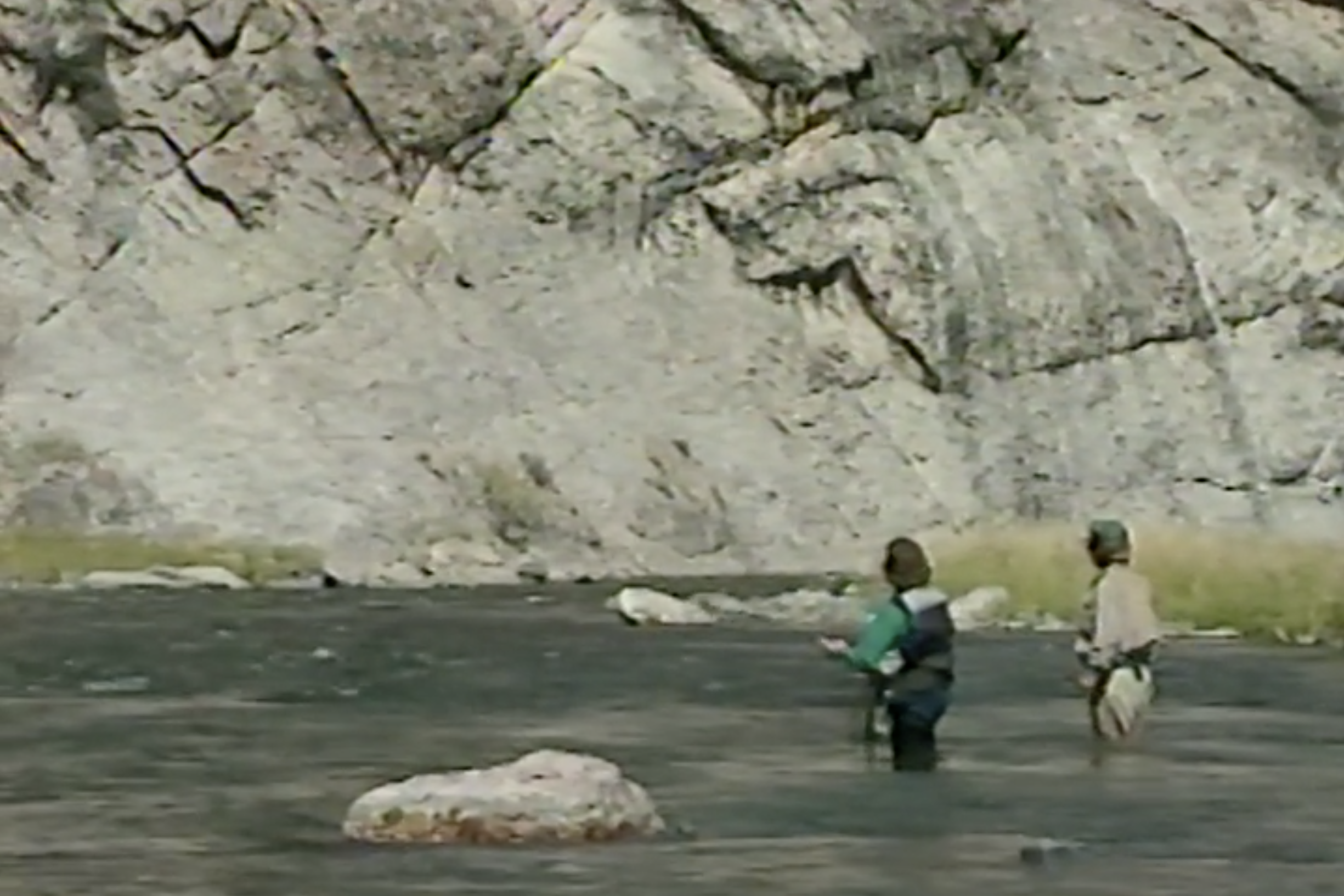 Two anglers, a woman and a man, stand just over knee-deep in a river. There is a large rock face in the background, indicating they are in a canyon.
