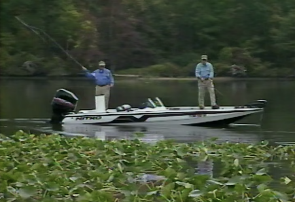 Two men stand on a motorboat. One is in the process of casting, while the other appears to be retrieving line.