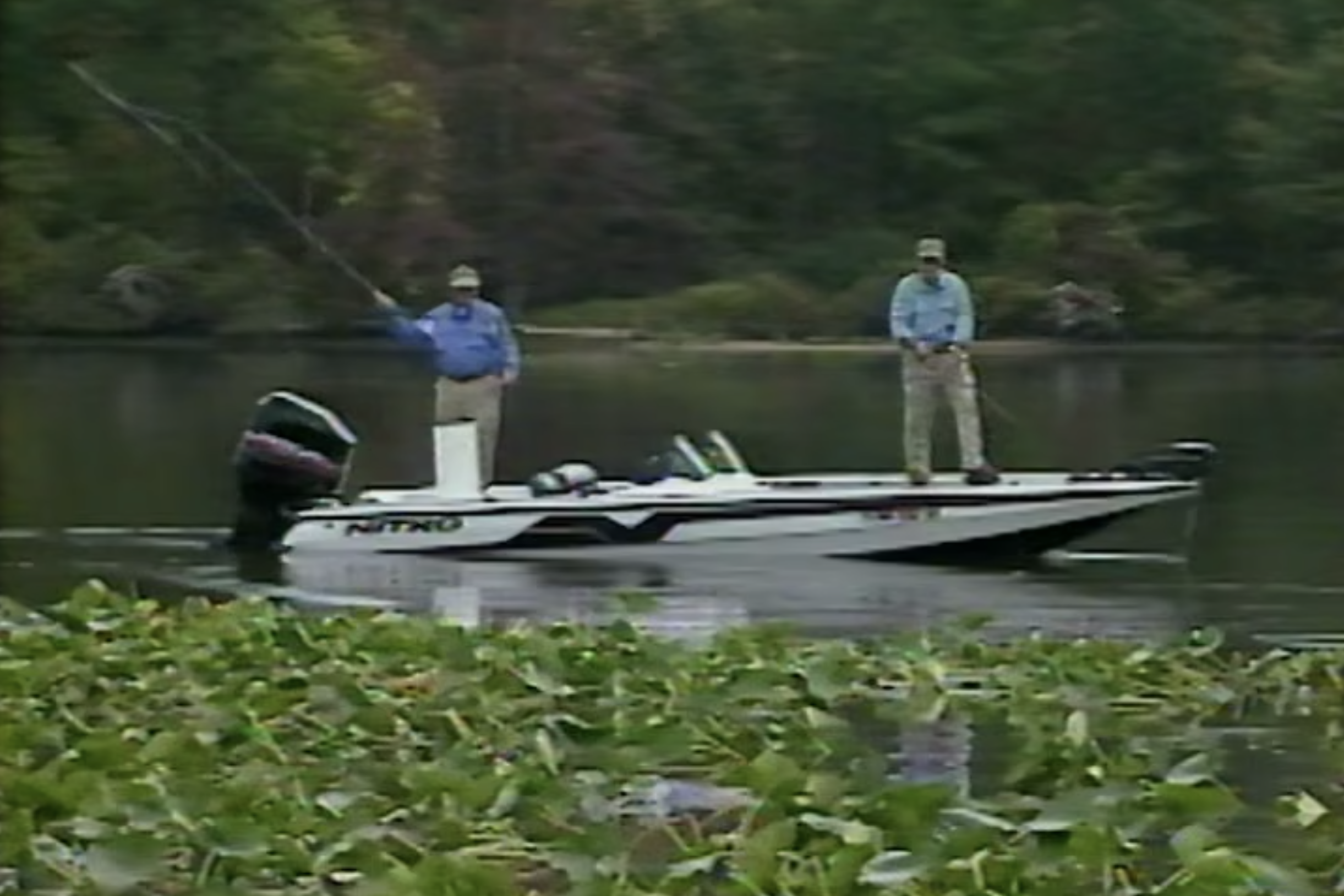 Two men stand on a motorboat. One is in the process of casting, while the other appears to be retrieving line.